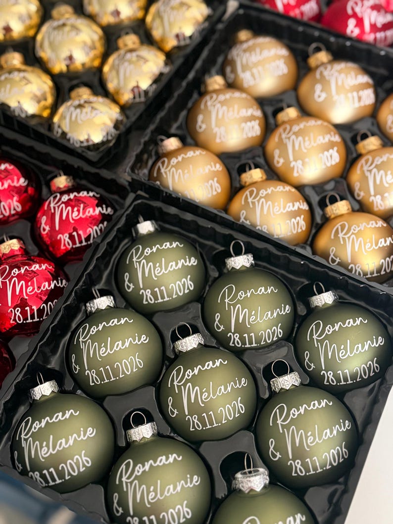K&ouml;nnte beinhalten: Eine Sammlung runder Weihnachtskugeln in mattem Olivgr&uuml;n, Metallic-Gold und gl&auml;nzendem Rot. Jede Kugel ist mit dem wei&szlig;en Schreibschrift-Text "Ronan & M&eacute;lanie" und dem Datum "28.11.2026" auf der Vorderseite bedruckt.