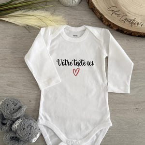 Peut inclure: Body de bébé blanc à manches longues. Le texte "Votre texte ici" est imprimé sur le devant, avec un cœur rouge. Des chaussons ours gris sont à côté du body. Un petit pot de plante et une tranche de bois sont en arrière-plan.