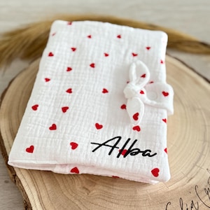 Könnte beinhalten: Ein weißes Musselin-Spucktuch für Babys mit rotem Herzmuster und dem Namen "Alba" in schwarzer Stickerei. Das Tuch hat eine geknotete Schleife und einen Druckknopfverschluss.