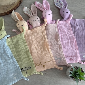 May include: A collection of pastel-colored baby security blankets with bunny heads. Each blanket features a different color, including blue, green, peach, pink, and purple. Some blankets have embroidered names and small designs.