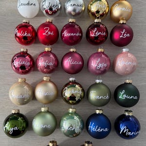 K&ouml;nnte beinhalten: Eine Sammlung von siebenundzwanzig runden Weihnachtskugeln in verschiedenen Farben wie Wei&szlig;, Silber, Gold, Rot, Rosa, Gr&uuml;n, Blau und Braun. Jede Kugel ist auf der Vorderseite mit einem individuellen Namen in wei&szlig;er Schreibschrift versehen.