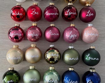 Puede incluir: Una colección de veintisiete adornos navideños redondos en varios colores, incluyendo blanco, plata, oro, rojo, rosa, verde, azul y marrón. Cada adorno presenta un nombre único escrito en letra cursiva blanca en la superficie frontal.