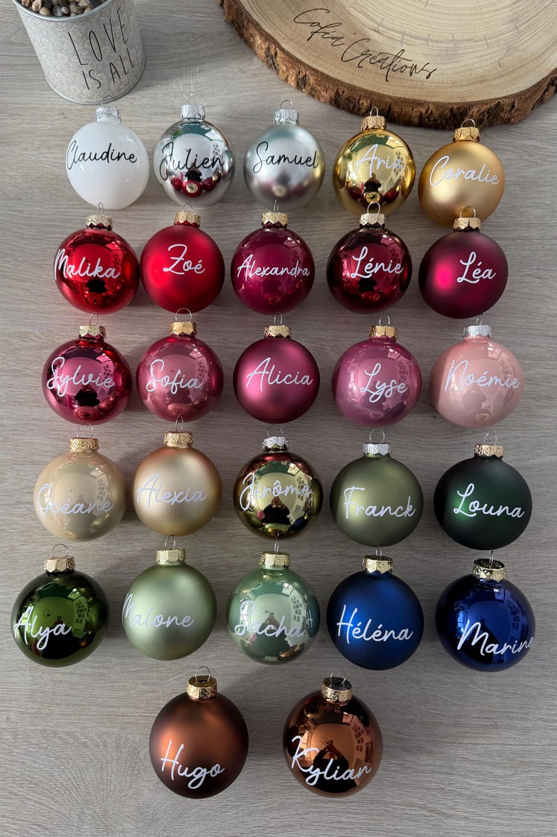K&ouml;nnte beinhalten: Eine Sammlung von siebenundzwanzig runden Weihnachtskugeln in verschiedenen Farben wie Wei&szlig;, Silber, Gold, Rot, Rosa, Gr&uuml;n, Blau und Braun. Jede Kugel ist auf der Vorderseite mit einem individuellen Namen in wei&szlig;er Schreibschrift versehen.