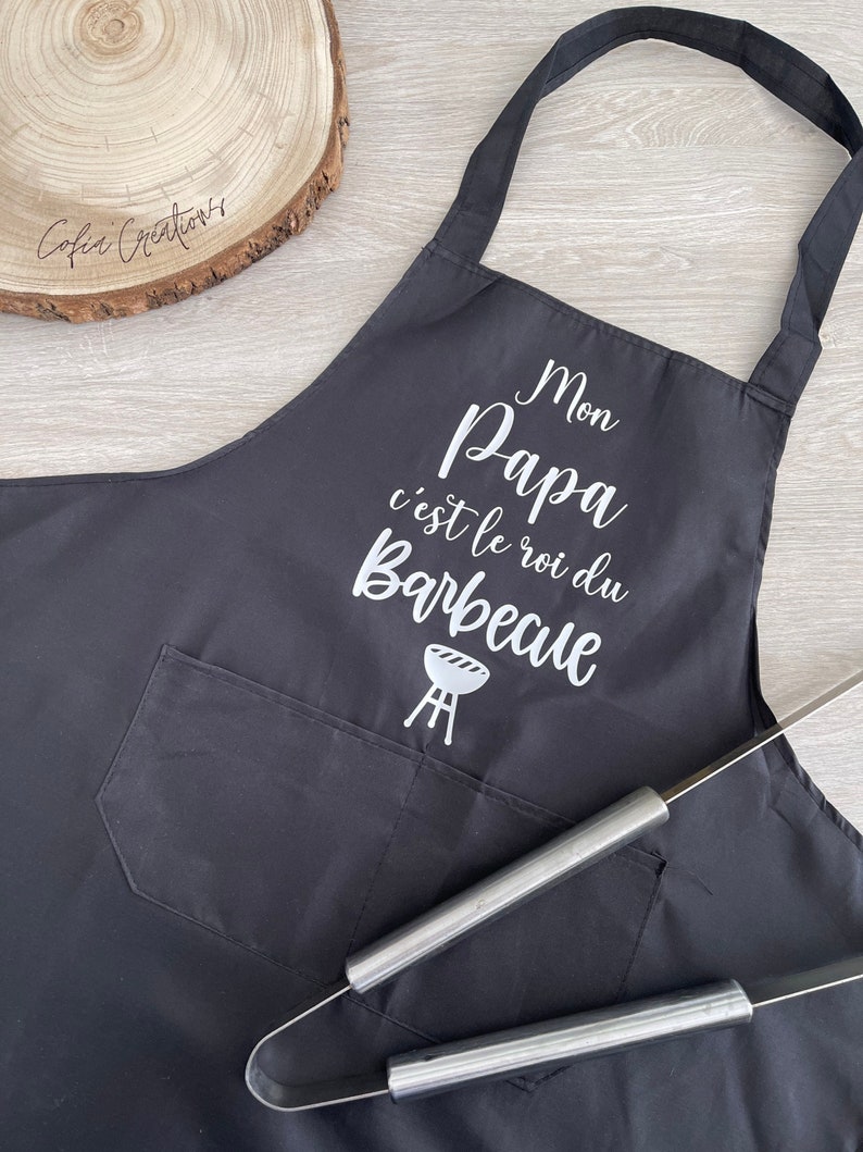May include: Black apron with a white pocket and the text "Mon Papa c'est le roi du Barbecue" with a grill icon.
