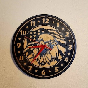 May include: A black and wood clock with a bald eagle design. The eagle is centered on the clock face with the American flag behind it. The clock face has black numbers and a red minute hand and black hour hand.