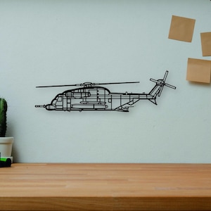 May include: Black metal helicopter wall art. The artwork is mounted on a white wall above a wooden desk. Other items on the desk include books, a camera, a small cactus, and a wooden artist's mannequin.