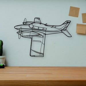 May include: A black metal airplane wall art piece with propeller details. The decorative item is mounted on a white wall above a wooden desk. A camera, books, and a cactus are on the desk.