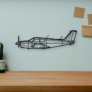 May include: A black metal airplane wall art piece is mounted on a white wall. The art is a wireframe outline of a small aircraft. A wooden desk in the foreground holds books, a camera, a cactus, and a wooden artist's mannequin.
