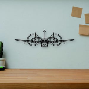 Puede incluir: Una pieza de arte mural de avión de metal negro con dos hélices, montada en una pared blanca. La obra de arte se exhibe sobre un escritorio de madera, con libros, una cámara y un cactus visibles.