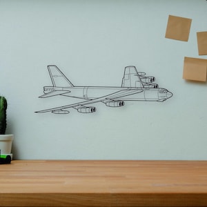 May include: A black wireframe illustration of a B-52 Stratofortress airplane is mounted on a white wall. A wooden desk holds a camera, books, a cactus, and a wooden artist's mannequin. A vase with branches is on the right.