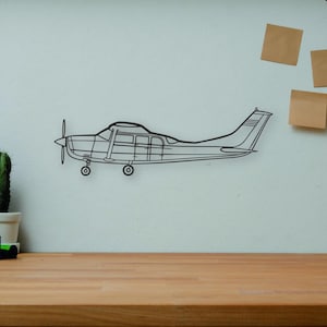 May include: A wireframe airplane wall art piece is mounted on a light-colored wall. The image also includes a vintage camera, books, a small cactus, a wooden artist's mannequin, and a decorative vase. The scene is set on a wooden desk.