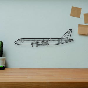 May include: A black metal airplane wall art piece is mounted on a white wall. The image also includes a wooden desk, books, a camera, a cactus, a wooden artist's mannequin, and a decorative vase.