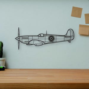 May include: A black metal airplane wall art piece with a propeller is mounted on a light blue wall. A wooden desk holds books, a camera, a cactus, and a wooden artist's figure. Decorative items.