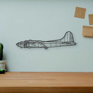Puede incluir: Una pieza de arte mural de avión de alambre negro está montada en una pared de color claro. Un escritorio de madera en primer plano contiene una cámara, libros, un cactus y un maniquí de artista de madera. También se ve un jarrón decorativo y tres notas adhesivas.