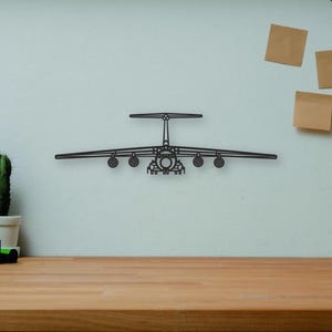 May include: A black metal airplane wall art piece. The airplane has a detailed design with wings, a tail, and multiple engines. The art is mounted on a light-colored wall above a wooden desk. Other items on the desk include books, a camera, and a cactus.