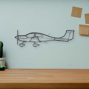 May include: A black metal airplane wall art piece is mounted on a white wall. A wooden desk in the foreground holds books, a camera, a cactus, and a wooden artist's mannequin. Decorative items include a vase and three square sticky notes.