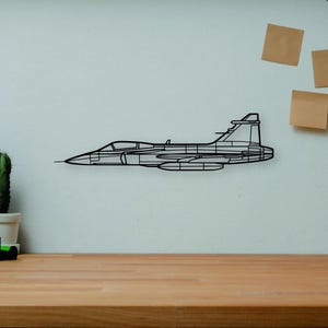 May include: A black metal wireframe of a jet airplane is mounted on a white wall. The jet is in profile, showing the fuselage, wings, and tail. A wooden desk in the foreground holds books, a camera, a cactus, and a wooden artist's mannequin.