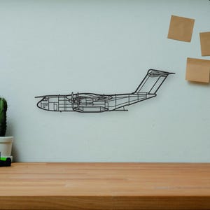 Peut inclure: Une décoration murale en forme d'avion en fil noir est accrochée à un mur clair. Un bureau en bois accueille un appareil photo, des livres, un cactus et un mannequin d'artiste en bois. Un vase décoratif et trois post-it sont également visibles.
