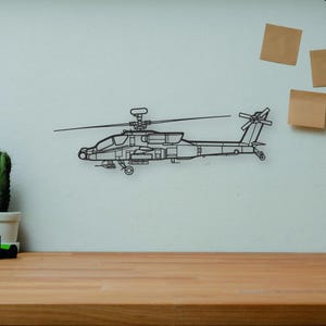 May include: A wireframe outline of a helicopter is mounted on a white wall. A wooden desk in the foreground holds a camera, books, a cactus, and a wooden artist's mannequin. Decorative elements include a vase and sticky notes.