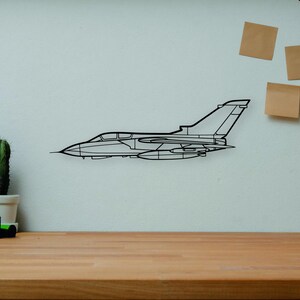 May include: A black metal outline of a jet airplane is mounted on a white wall. The jet is above a wooden desk with a camera, books, a cactus, and a wooden artist's mannequin. A decorative vase and three sticky notes are also on the wall.