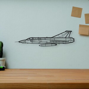May include: A black wireframe airplane wall art piece is mounted on a white wall above a wooden desk. A camera, books, and a cactus are on the desk. A wooden artist's mannequin and a vase are also present.