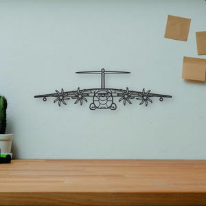 Peut inclure: Une décoration murale en métal noir en forme d'avion avec un design d'hélice est fixée sur un mur blanc. L'avion est centré, avec un bureau en bois au premier plan. Un appareil photo, des livres et un cactus sont sur le bureau.