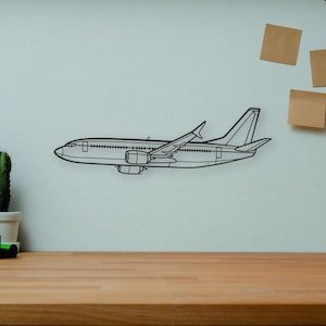 Puede incluir: En un escritorio de madera hay una cámara negra y plateada, libros, un cactus en una maceta blanca y un coche de juguete. Un dibujo lineal negro de un avión está en la pared. También hay un maniquí de artista de madera y un jarrón decorativo.