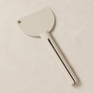 Puede incluir: Un raspador de cocina de color beige claro con una hoja semicircular y un mango largo y acanalado. La hoja presenta un pequeño orificio circular cerca de la esquina superior izquierda para colgar, y el mango tiene una ranura vertical.