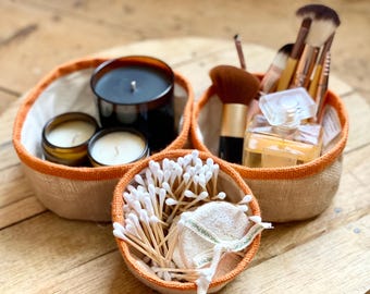 Puede incluir: Tres pequeñas cestas redondas tejidas con ribete naranja. Una contiene velas, otra brochas de maquillaje y perfume, y la tercera bastoncillos de algodón y una esponja vegetal. Sobre una superficie de madera.