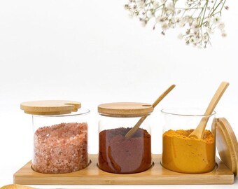 Puede incluir: Un especiero de bambú con tres frascos de vidrio transparente, cada uno con una especia diferente. Los frascos tienen tapas y cucharas de madera. Un frasco contiene sal rosa, otro especia roja y el tercero especia amarilla. Una cuchara de madera descansa delante.