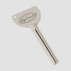 May include: A light beige tube squeezer with the text "UpCircle" in a black oval. The squeezer has a semi-circular head with a hole and a ribbed handle. The handle has a slot for inserting a tube.