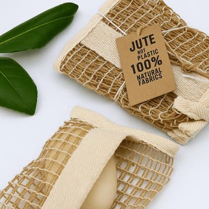 May include: Two beige and cream-colored jute net bags with a tag that reads "JUTE NOT PLASTIC 100% NATURAL FABRICS". The bags are made of woven jute and cream-colored fabric. Green leaves are in the background.