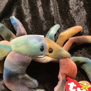 Puede incluir: Un cangrejo Ty Beanie Baby multicolor con la cara blanca y los ojos negros. El cangrejo tiene una etiqueta Ty roja y blanca unida a una de sus patas. La etiqueta dice "Beanie Original Babies".