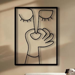 Puede incluir: Arte mural de metal negro que presenta un dibujo lineal minimalista de una cara con los ojos cerrados y una mano tocando la nariz. La obra de arte está enmarcada en un borde negro y cuelga de una pared beige. La habitación también incluye un sofá blanco y un perro.