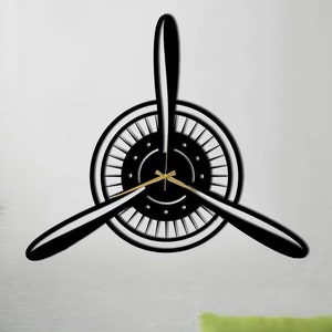May include: Black metal wall clock shaped like an aeroplane propeller. The clock has a gold-coloured hand and is mounted on a white wall. A green sofa with a throw pillow is in the foreground.