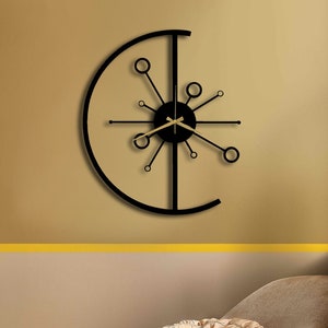 Könnte beinhalten: Eine moderne Wanduhr in Schwarz und Gold mit einem runden Design und einem Sternenausbruchmuster. Die Uhr hängt an einer beigefarbenen Wand über einem braunen Sessel.