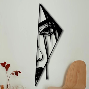 Può includere: Un'opera d'arte da parete in metallo nero raffigurante un volto stilizzato all'interno di una cornice triangolare. L'opera presenta un singolo occhio, un naso e una bocca, con linee a forma di lacrima. È montata su un muro bianco, con una sedia e un tavolo in legno in primo piano.