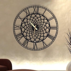 Op de afbeelding: Een grote zwarte metalen wandklok met Romeinse cijfers en een decoratief ontwerp van overlappende cirkels. De klok hangt aan een beige muur boven een bruine leren fauteuil en een voetenbank.