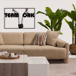 May include: A black metal wall art piece with the words "TEAM WORK" and a graphic of two figures pulling a weight. The art is mounted above a beige sofa with decorative pillows and a white coffee table. A potted plant is in the background.