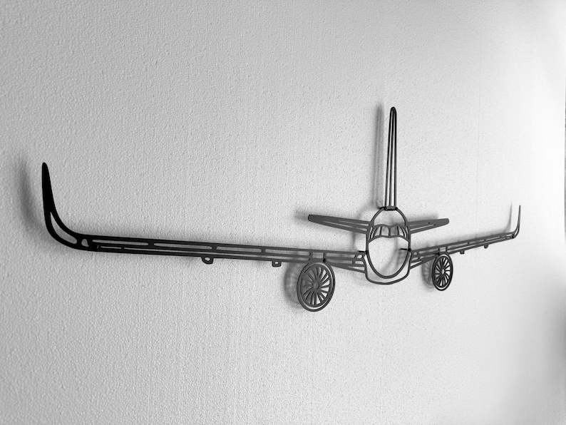 May include: A black metal airplane wall art piece. The artwork features a detailed outline of an airplane, including the fuselage, wings, tail, and wheels. The piece is mounted on a textured white wall, creating a striking contrast.