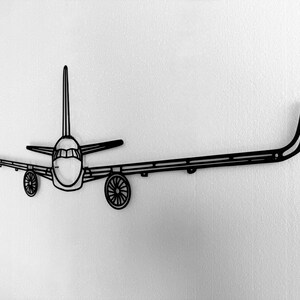 May include: Black metal airplane wall art. The artwork features a detailed outline of an airplane, including the fuselage, wings, tail, and wheels. The design is mounted on a white wall, creating a striking contrast.