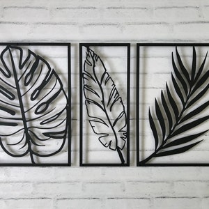 Puede incluir: Tres piezas de decoraci&oacute;n de pared de metal negro con dise&ntilde;os recortados de hojas tropicales. Las hojas son de estilo minimalista y est&aacute;n enmarcadas en metal negro.
