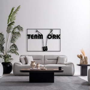 May include: Black metal wall art spelling "TEAM WORK" with a stylized design of people pulling a rope. The artwork is mounted on a white wall above a gray sofa, with a black and brown coffee table and a large potted plant.