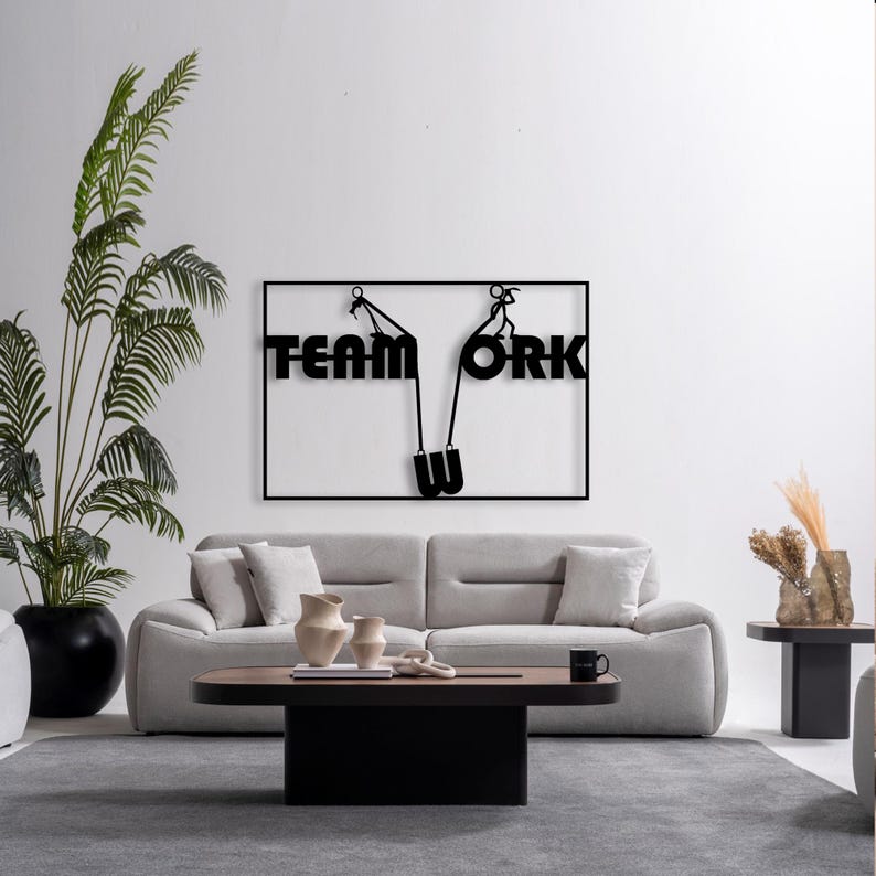 May include: Black metal wall art spelling "TEAM WORK" with a stylized design of people pulling a rope. The artwork is mounted on a white wall above a gray sofa, with a black and brown coffee table and a large potted plant.