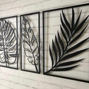 Puede incluir: Tres piezas de decoraci&oacute;n de pared de metal negro con dise&ntilde;os recortados de hojas tropicales. Las hojas son de estilo minimalista y est&aacute;n dispuestas en fila.