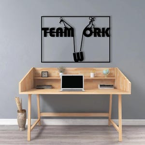 May include: A wooden desk with a laptop, books, and decorative items. Above the desk is a black metal sign that reads "TEAM WORK" with a graphic of people pulling up the letters. The desk has a natural wood finish.