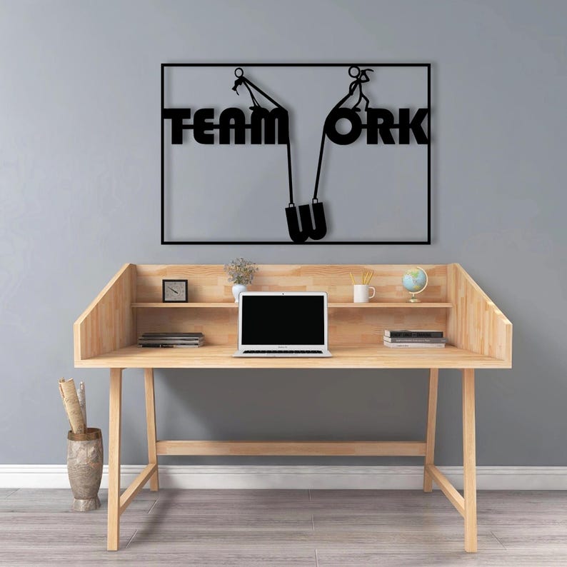 May include: A wooden desk with a laptop, books, and decorative items. Above the desk is a black metal sign that reads "TEAM WORK" with a graphic of people pulling up the letters. The desk has a natural wood finish.
