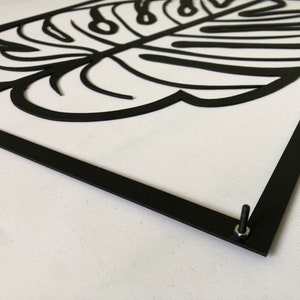 Puede incluir: Decoraci&oacute;n de pared de metal negro con un gran recorte detallado de una hoja de monstera. La hoja est&aacute; enmarcada con un borde de metal negro y tiene un tornillo en la esquina inferior derecha.