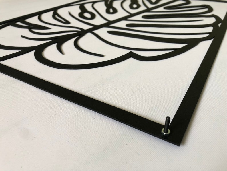 Puede incluir: Decoraci&oacute;n de pared de metal negro con un gran recorte detallado de una hoja de monstera. La hoja est&aacute; enmarcada con un borde de metal negro y tiene un tornillo en la esquina inferior derecha.