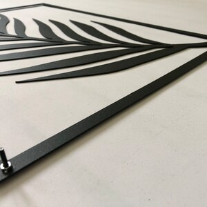 Puede incluir: Arte de pared de metal negro con un dise&ntilde;o estilizado de hoja de palma. El arte est&aacute; enmarcado con un marco de metal negro y tiene dos tornillos en la parte inferior.